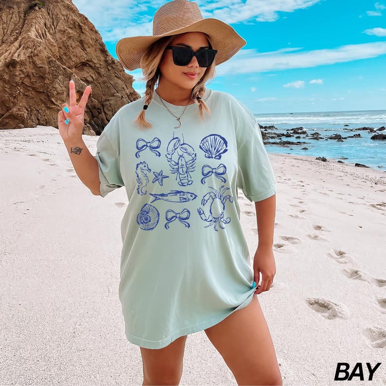May include: A light blue t-shirt with a graphic print of various sea creatures, including a crab, a seahorse, a fish, a seashell, and a lobster. The shirt has a relaxed fit and a crew neckline.