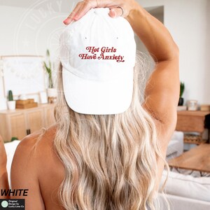 May include: White baseball cap with the text "Hot Girls Have Anxiety" in red script. The cap is made of a soft material and has a curved brim. The cap is being worn by a person with blonde hair.