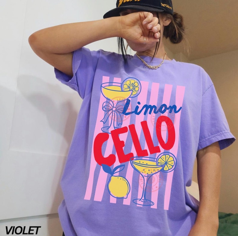 May include: Violet-colored t-shirt with a graphic design featuring two cocktail glasses, lemons, and the words "Limon Cello" in red and blue. The design is set against a pink and white striped background. The word "VIOLET" is printed in white at the bottom left.