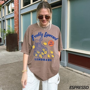 Puede incluir: Una camiseta marr&oacute;n con un gr&aacute;fico de una bolsa de limones y el texto "Freshly Squeezed 100% Natural Lemonade".