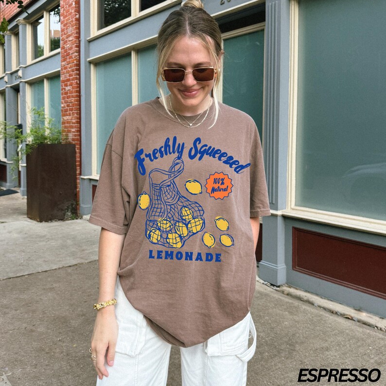 Puede incluir: Una camiseta marr&oacute;n con un gr&aacute;fico de una bolsa de limones y el texto "Freshly Squeezed 100% Natural Lemonade".