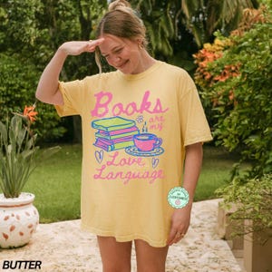 以下が含まれることがあります： ピンクと青で「Books are my Love Language」と書かれた黄色のTシャツ。グラフィックには、本の山、ティーカップ、ハートが含まれています。 ゆったりとしたオーバーサイズのフィット感のシャツです。