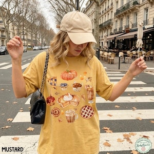 May include: A mustard yellow t-shirt featuring a graphic print of various autumn-themed treats including a pumpkin, pies, cinnamon rolls, and lattes. The shirt is paired with a beige baseball cap and a black quilted shoulder bag with a chain strap.