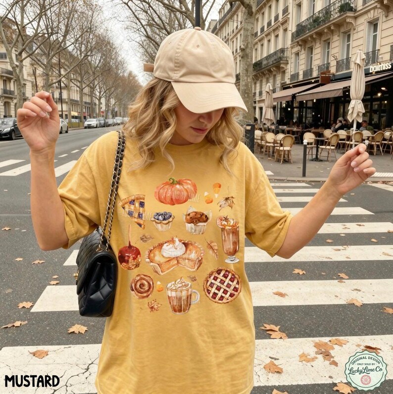 May include: A mustard yellow t-shirt featuring a graphic print of various autumn-themed treats including a pumpkin, pies, cinnamon rolls, and lattes. The shirt is paired with a beige baseball cap and a black quilted shoulder bag with a chain strap.