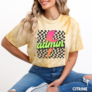 May include: A yellow tie-dye t-shirt with a black and white chequered pattern and a pink and green lightning bolt graphic with the word "admin" in green text.