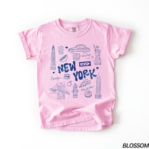 May include: Pink t-shirt with a blue graphic of New York City landmarks, including the Statue of Liberty, the Empire State Building, and the Brooklyn Bridge. The text "New York" is prominently displayed.