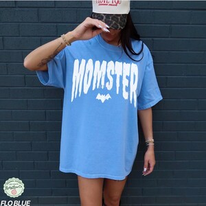 May include: A light blue t-shirt with the word "MOMSTER" in large white letters and a bat graphic. The shirt is a relaxed fit and has short sleeves. The text "FLO BLUE" is at the bottom of the image.