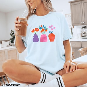 May include: A light blue t-shirt with a graphic of three colorful vases with flowers. The vases are purple, pink, and orange. The flowers are red, orange, yellow, blue, and green.