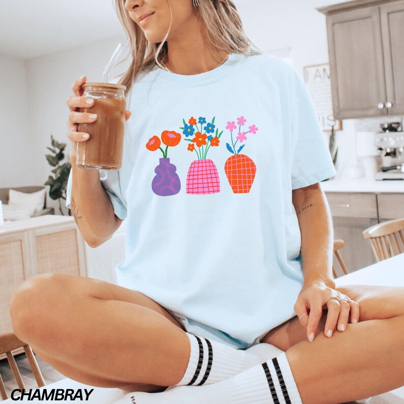 May include: A light blue t-shirt with a graphic of three colorful vases with flowers. The vases are purple, pink, and orange. The flowers are red, orange, yellow, blue, and green.