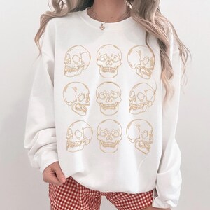 May include: A white crewneck sweatshirt featuring a repeating grid pattern of nine gold-colored line art skulls. The graphic consists of three rows of three skulls, alternating between front-facing and side-profile views, printed on the center of the chest.