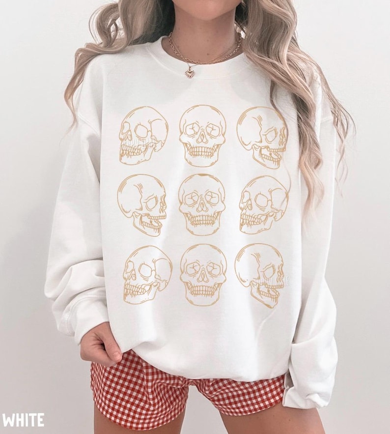 May include: A white crewneck sweatshirt featuring a repeating grid pattern of nine gold-colored line art skulls. The graphic consists of three rows of three skulls, alternating between front-facing and side-profile views, printed on the center of the chest.