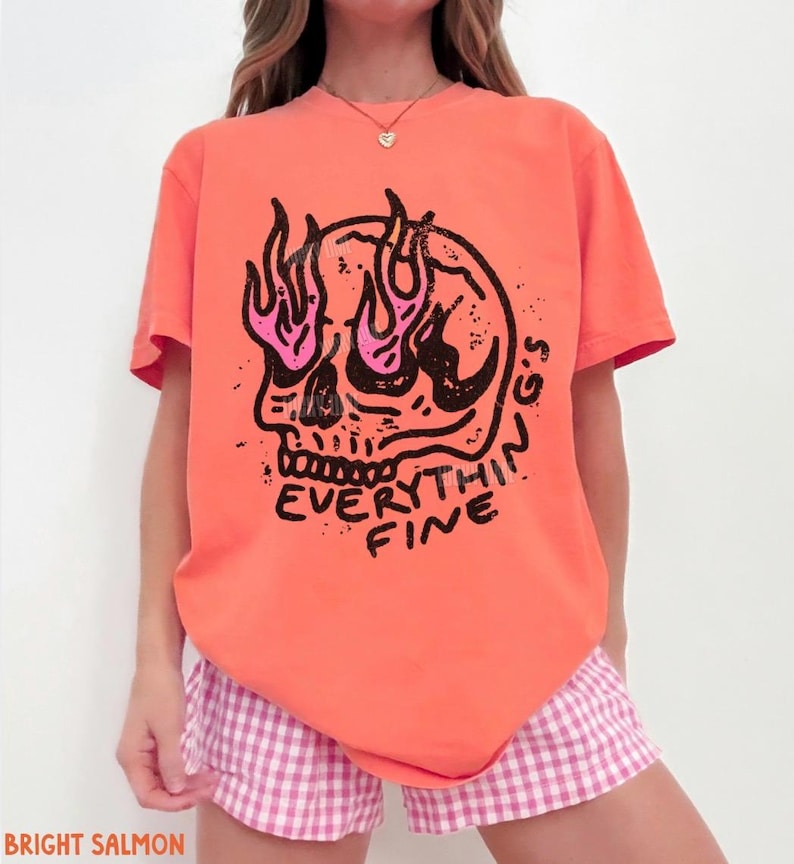 May include: A bright salmon t-shirt featuring a black graphic of a skull with pink flames emanating from the eye sockets. Below the skull, the text reads "EVERYTHING'S FINE" in a bold, hand-drawn style font. The shirt has short sleeves and a crew neck.