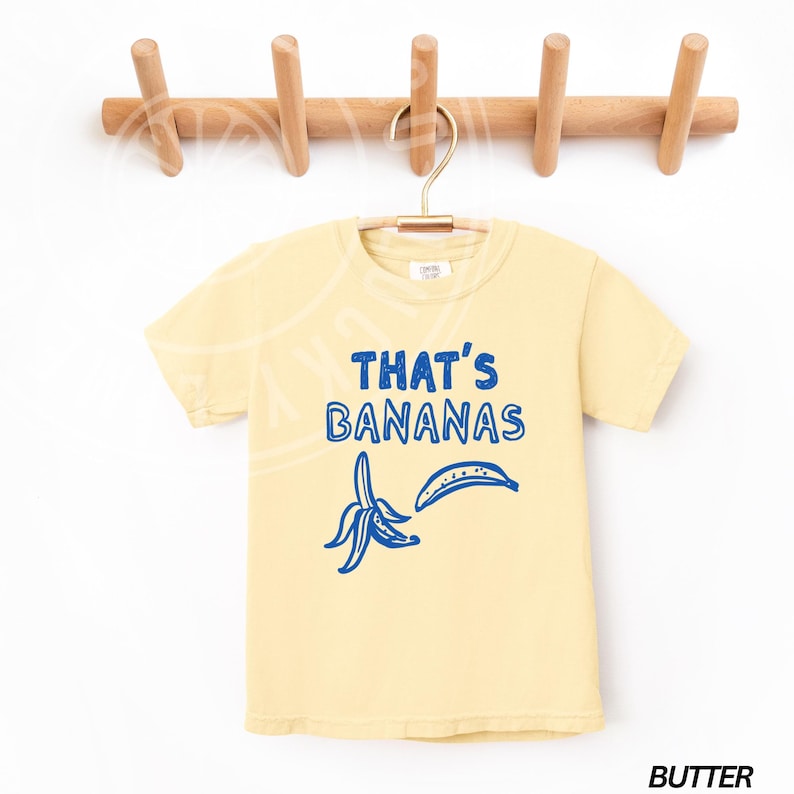 Op de afbeelding: Een geel T-shirt met een blauwe afbeelding van een banaan en de tekst "THAT'S BANANAS".
