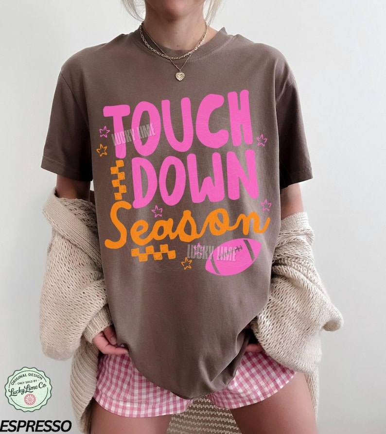 May include: Espresso-colored t-shirt with the words "TOUCH DOWN Season" in pink and orange, along with a pink football graphic. The shirt has a relaxed fit and is worn with a beige cardigan and pink gingham shorts. The design includes the text "LUCKY LIME".