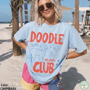 Puede incluir: Camiseta azul claro con las palabras "DOODLE mom CLUB" en naranja. La camiseta presenta ilustraciones de dos caniches y la frase "life is better with doodles". La camiseta forma parte de la colecci&oacute;n "Lucky Lane Co".