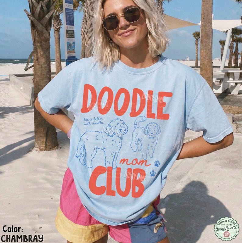 Puede incluir: Camiseta azul claro con las palabras "DOODLE mom CLUB" en naranja. La camiseta presenta ilustraciones de dos caniches y la frase "life is better with doodles". La camiseta forma parte de la colecci&oacute;n "Lucky Lane Co".