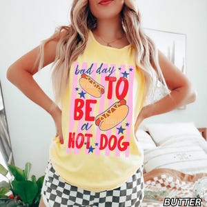 Op de afbeelding: Geel mouwloos shirt met de tekst "bad day to be a hot dog" in rood en blauw. De afbeelding bevat twee hotdogs en blauwe sterren op een roze gestreepte achtergrond.