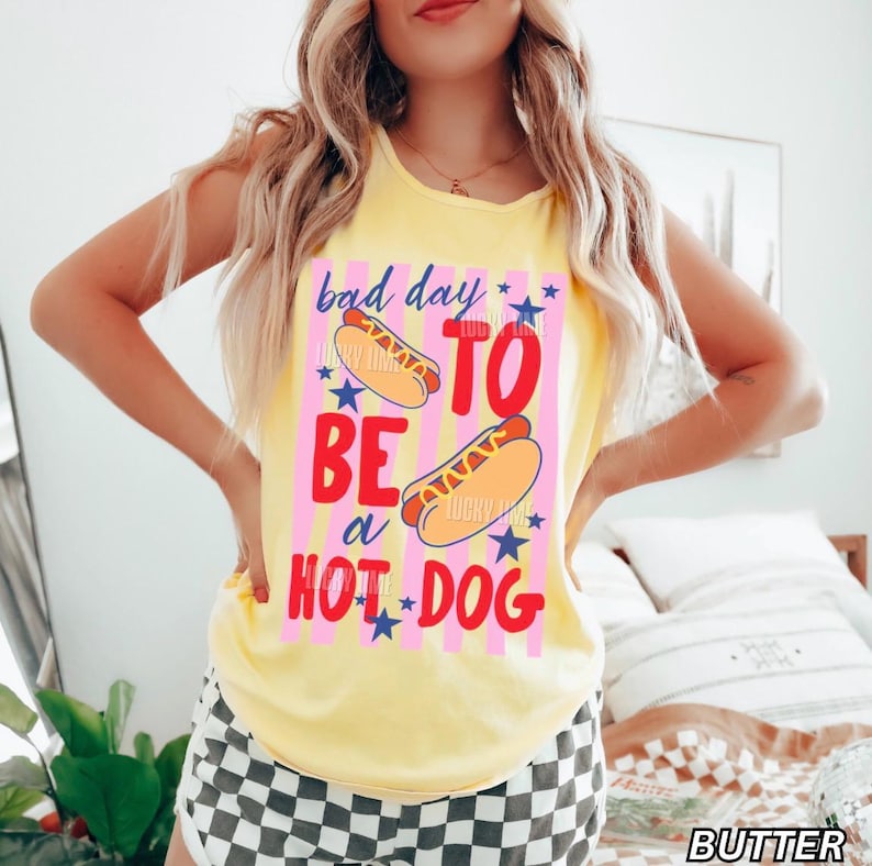 Op de afbeelding: Geel mouwloos shirt met de tekst "bad day to be a hot dog" in rood en blauw. De afbeelding bevat twee hotdogs en blauwe sterren op een roze gestreepte achtergrond.