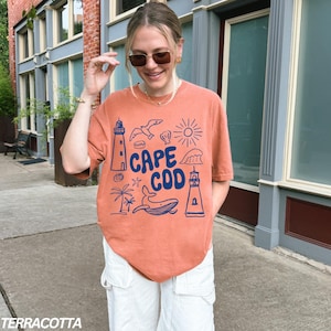 Peut inclure: Un t-shirt orange terre cuite avec un motif graphique bleu qui dit "Cape Cod" et repr&eacute;sente un phare, une baleine, une coquille, un palmier et un soleil.