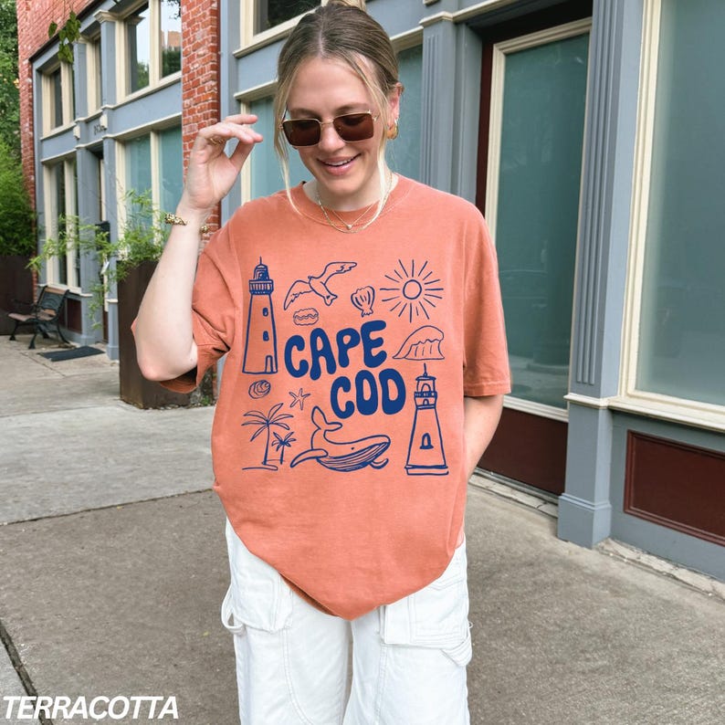Peut inclure: Un t-shirt orange terre cuite avec un motif graphique bleu qui dit "Cape Cod" et repr&eacute;sente un phare, une baleine, une coquille, un palmier et un soleil.