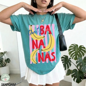Puede incluir: Camiseta verde mar con la frase "that's BANANAS" en un dise&ntilde;o l&uacute;dico. El gr&aacute;fico presenta pl&aacute;tanos amarillos y rayas verticales rosas y azules. La camiseta es de estilo casual y c&oacute;modo.