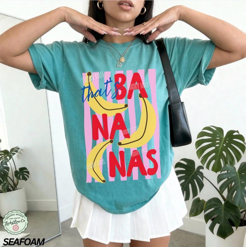 Puede incluir: Camiseta verde mar con la frase "that's BANANAS" en un dise&ntilde;o l&uacute;dico. El gr&aacute;fico presenta pl&aacute;tanos amarillos y rayas verticales rosas y azules. La camiseta es de estilo casual y c&oacute;modo.