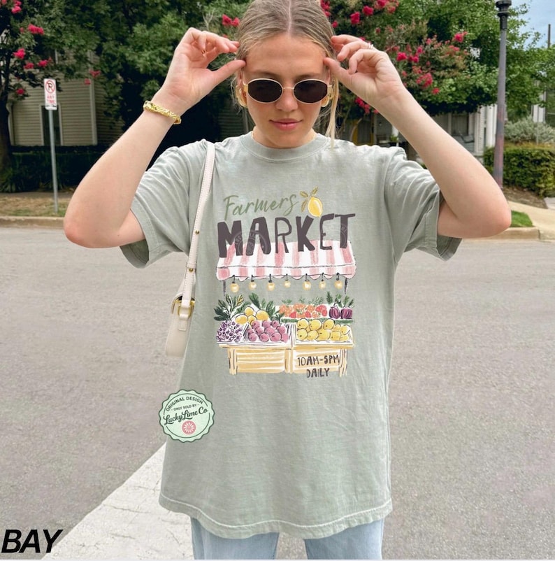 May include: Sage green t-shirt with a "Farmers Market" graphic. The design features a market stall with various fruits and vegetables. The text "10AM-5PM DAILY" is also visible. The shirt has a relaxed fit.