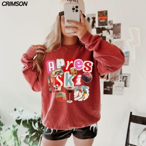 May include: A red-orange sweatshirt with the words "Apres Ski" in a colorful collage design. The design includes images of ski goggles, a camera, cocktails, a snow globe, and skiers. The word "CRIMSON" is in the top left corner.