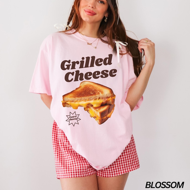 May include: A pink t-shirt with a graphic of a grilled cheese sandwich and the text "Grilled Cheese" in brown letters. The sandwich is labeled "Extra Cheesy".