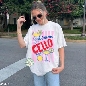 May include: White t-shirt with a colorful "Limon Cello" graphic. The design features cocktail glasses, lemons, and pink and white stripes. The shirt has a relaxed fit and short sleeves.