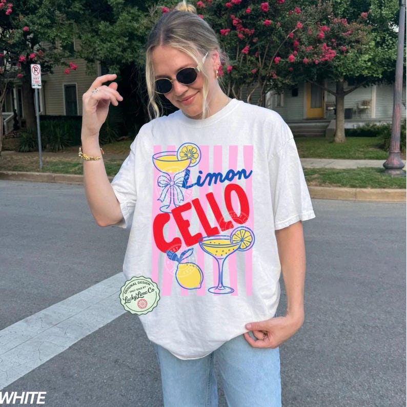 May include: White t-shirt with a colorful "Limon Cello" graphic. The design features cocktail glasses, lemons, and pink and white stripes. The shirt has a relaxed fit and short sleeves.