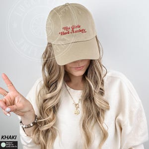 May include: Khaki baseball cap with the embroidered text "Hot Girls Have Anxiety" in red. The cap has a curved brim and a classic six-panel design. The hat is made of a soft, durable fabric.