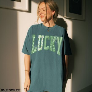 May include: Blue spruce t-shirt with the word "LUCKY" in green. The letters have various patterns, including checkered and striped designs. The shirt is a relaxed fit, with short sleeves and a crew neck.