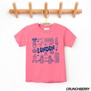 May include: Pink short-sleeved t-shirt with a blue graphic of London landmarks, including a double-decker bus, a red telephone box, Big Ben, and the words "LONDON!"