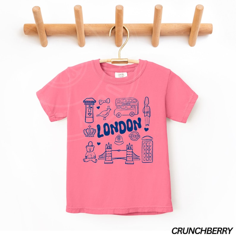 May include: Pink short-sleeved t-shirt with a blue graphic of London landmarks, including a double-decker bus, a red telephone box, Big Ben, and the words "LONDON!"
