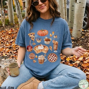 May include: A blue short-sleeve t-shirt featuring a graphic print of various autumn-themed treats including a pumpkin, a slice of pie, a muffin, a cinnamon roll, and mugs of coffee, all arranged in a cluster on the front of the garment.