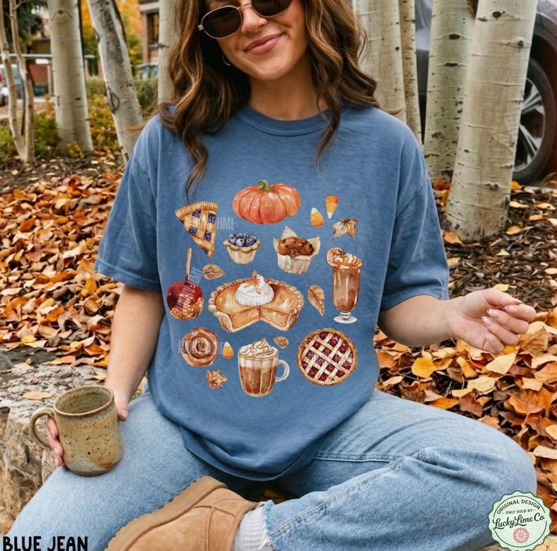 May include: A blue short-sleeve t-shirt featuring a graphic print of various autumn-themed treats including a pumpkin, a slice of pie, a muffin, a cinnamon roll, and mugs of coffee, all arranged in a cluster on the front of the garment.