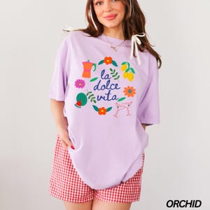 Pu&ograve; includere: Una maglietta color lavanda con una stampa grafica di un cerchio con il testo "la dolce vita" in blu e circondata da illustrazioni colorate di limoni, fiori, un'anguria, una caffettiera e bicchieri da martini.