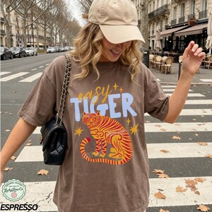 May include: Brown t-shirt with the text "easy TIGER" in blue and yellow, featuring a tiger illustration in red and yellow. The shirt is part of the "Lucky Lime Co" original design collection.