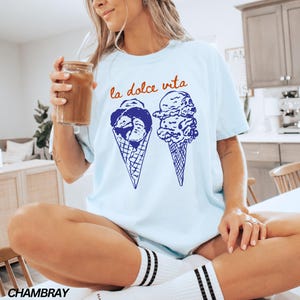 May include: A light blue t-shirt with the text "la dolce vita" in orange and blue. The shirt features a graphic of two ice cream cones with blue and white swirls.