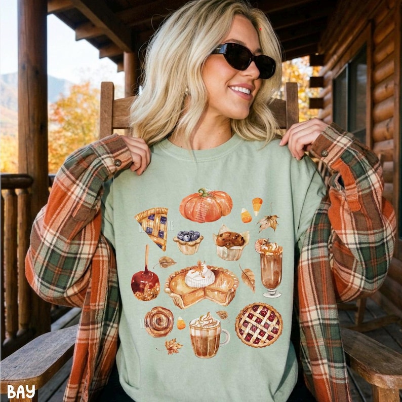 May include: A light green crewneck sweatshirt featuring a graphic print of various autumn-themed treats including a pumpkin, a slice of pie, a caramel apple, a cinnamon roll, a muffin, a latte, and a lattice-crust pie, worn under a plaid flannel shirt.