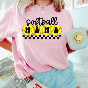 Op de afbeelding: Roze T-shirt met de tekst "Softball Mama" in zwarte letters met gele softball-afbeeldingen en een zwart-wit geruit patroon.