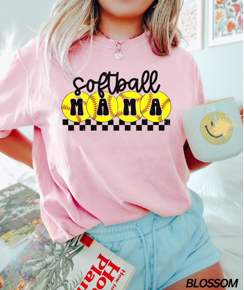 Op de afbeelding: Roze T-shirt met de tekst "Softball Mama" in zwarte letters met gele softball-afbeeldingen en een zwart-wit geruit patroon.