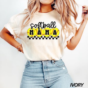 Op de afbeelding: Een wit T-shirt met de tekst "Softball Mama" in zwarte letters en vier gele softball-afbeeldingen. Het shirt wordt gedragen door een model dat blauwe jeans draagt.
