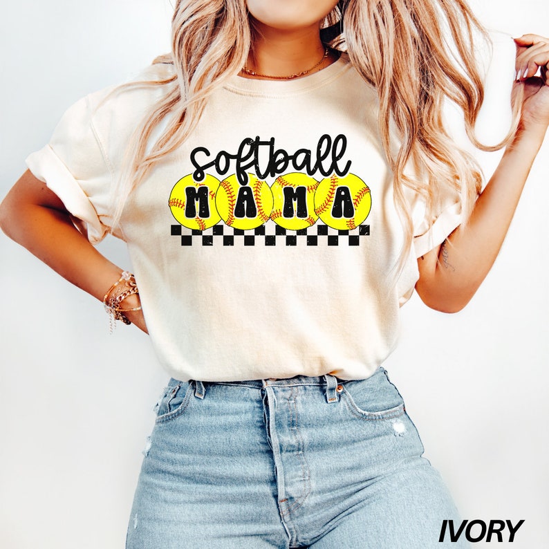 Op de afbeelding: Een wit T-shirt met de tekst "Softball Mama" in zwarte letters en vier gele softball-afbeeldingen. Het shirt wordt gedragen door een model dat blauwe jeans draagt.