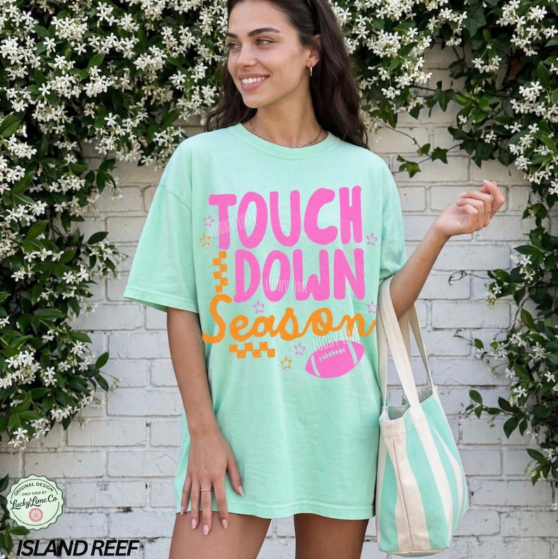 May include: A light green t-shirt with the words "TOUCH DOWN SEASON" in pink and orange, along with a pink football graphic. The shirt is paired with a striped tote bag.