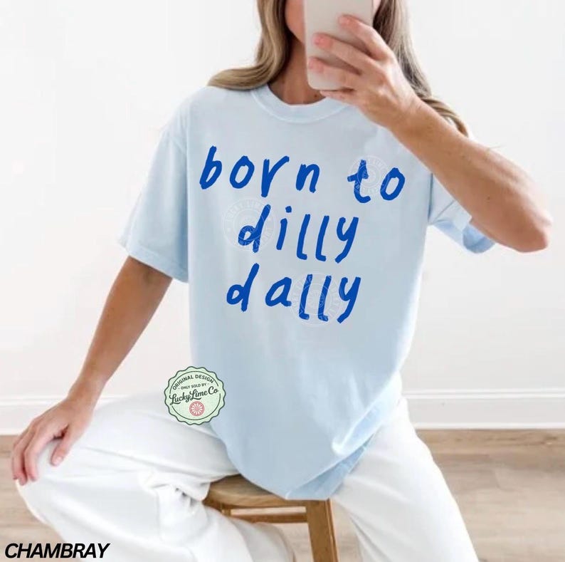 May include: Light blue t-shirt with the phrase "born to dilly dally" in blue lettering. The shirt has short sleeves and a crew neck. The shirt is part of the Lucky Lane Co. original design collection.