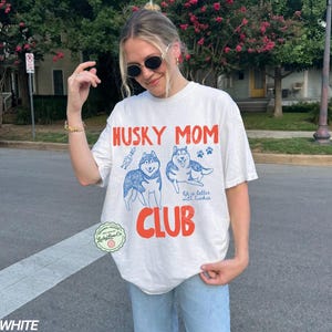 K&ouml;nnte beinhalten: Wei&szlig;es T-Shirt mit dem Text "HUSKY MOM CLUB" in Rot und Blau. Das Shirt zeigt Illustrationen von zwei Huskys und Pfotenabdr&uuml;cken. Der Satz "life is better with huskies" ist ebenfalls aufgedruckt.