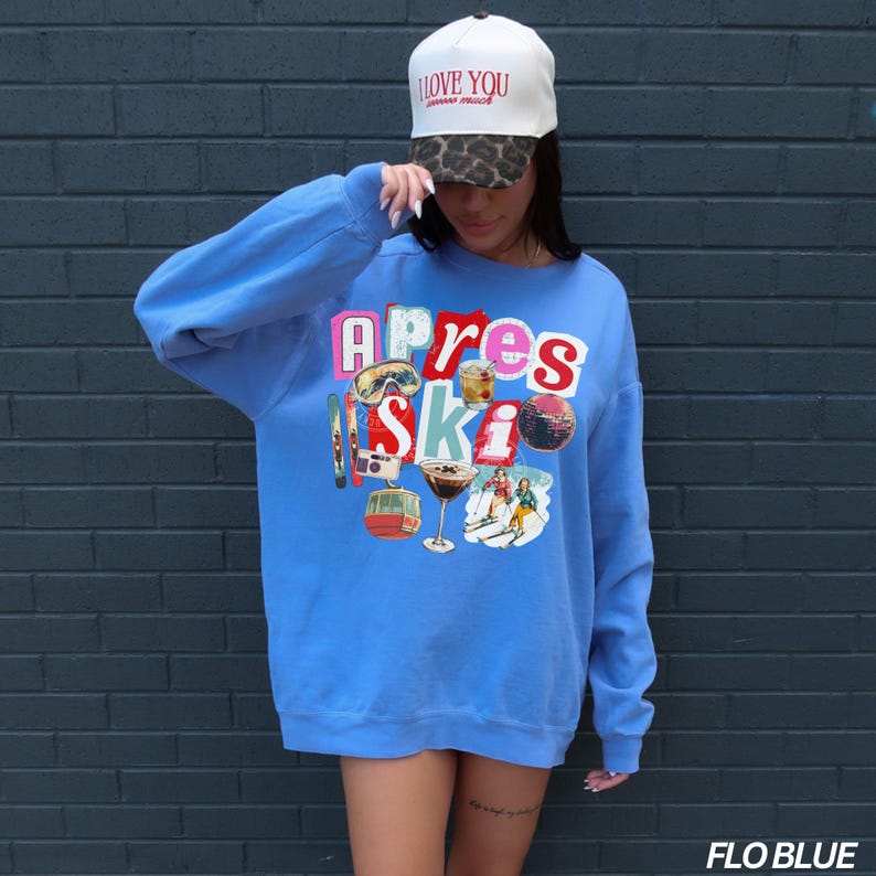 May include: A light blue sweatshirt with the words "Apres Ski" in a colorful, collage-style design. The design includes images of ski equipment, cocktails, and a ski lift. A white baseball cap with the text "I love you" is also visible.