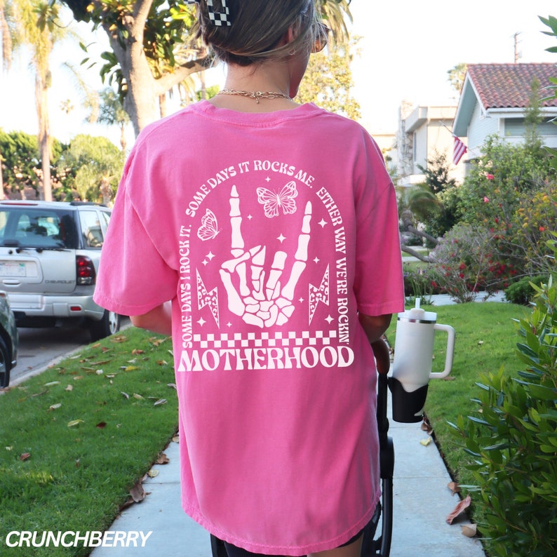 May include: A bright pink t-shirt with a white graphic on the back. The graphic includes a skeleton hand in a rock-and-roll gesture, butterflies, lightning bolts, and the word "MOTHERHOOD". The text "SOME DAYS I ROCK IT. EITHER WAY WE'RE ROCKIN'" is also included.